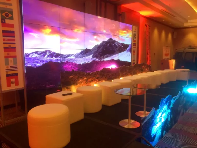 Un escenario de conferencias con asientos blancos tipo otomana, pequeñas mesas redondas y una gran pantalla que muestra un paisaje montañoso; a un lado se ven varias banderas de países.