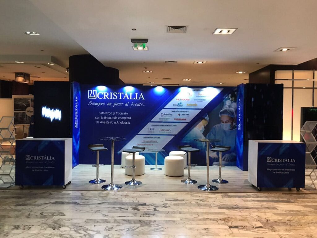 Stand de Cristália con sillas, mesas y expositores de marca en un centro de convenciones; el fondo muestra imágenes médicas y logotipos de patrocinadores.