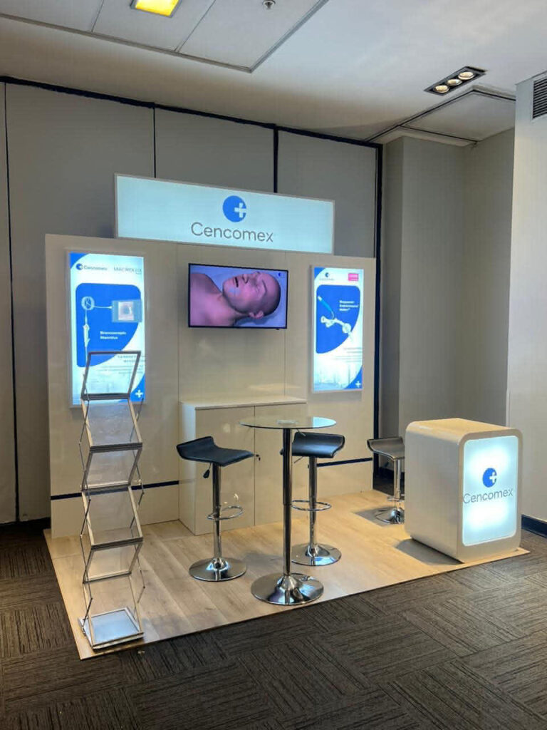 Stand de exposición para Cencomex con una pantalla de visualización, imágenes de productos, un soporte para folletos, dos sillas en una mesa redonda y un mostrador iluminado con el logotipo de la empresa.