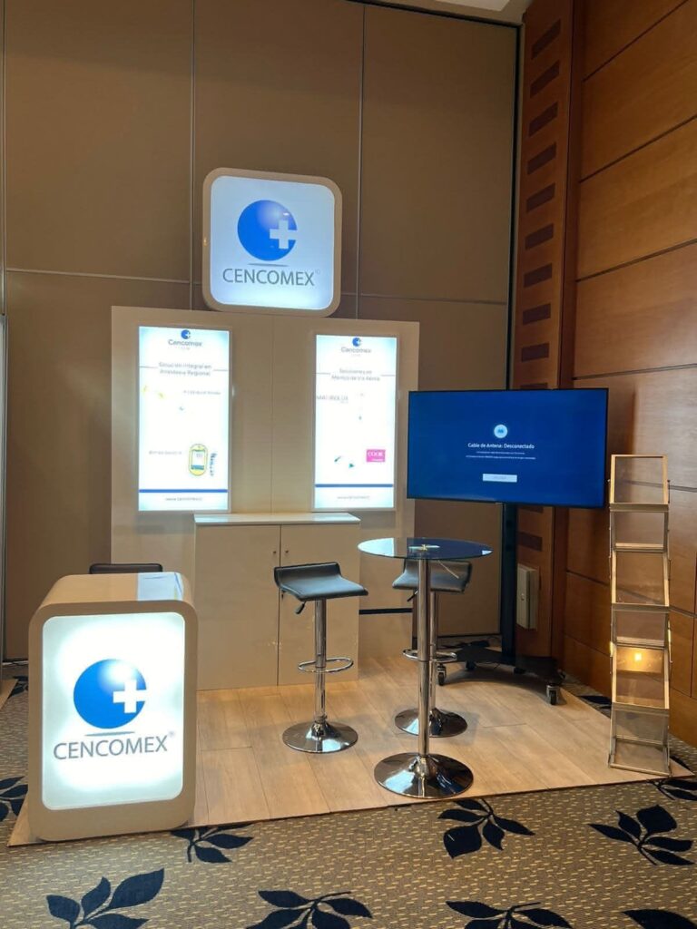 Stand de CENCOMEX con carteles luminosos, dos taburetes junto a una mesa redonda, un mostrador, una pantalla de TV y una estantería de madera, instalado en una sala de conferencias.