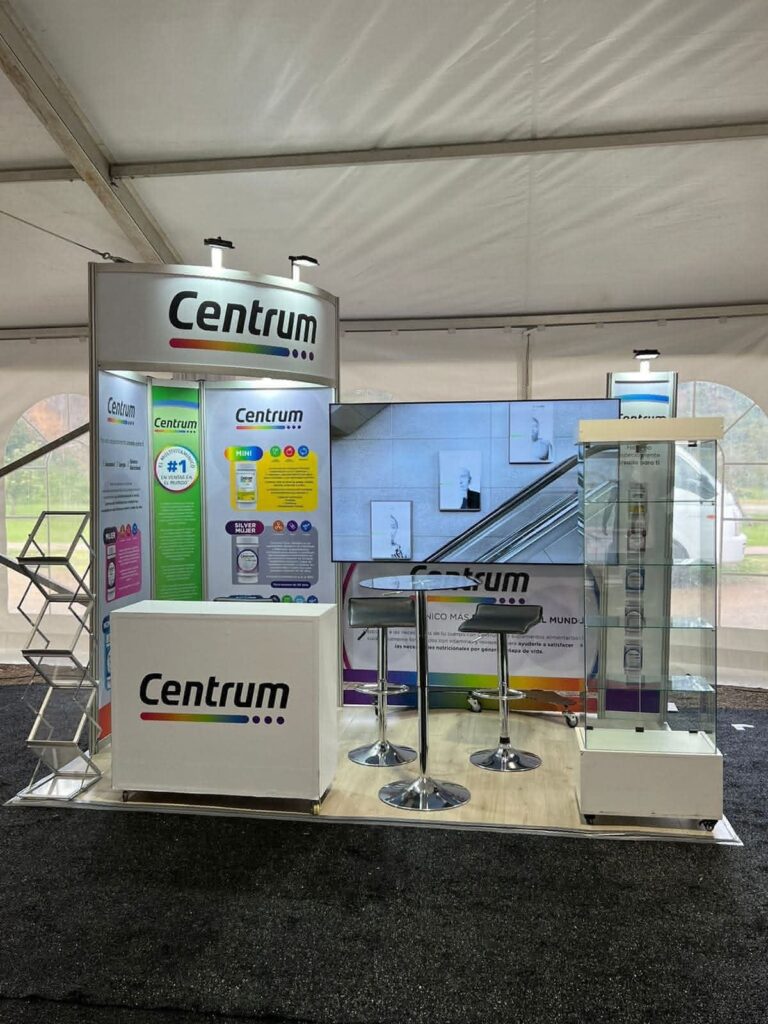 Stand promocional de Centrum con pancartas de la marca, expositores informativos, taburetes, un mostrador y una vitrina de cristal en el interior de una carpa para eventos.