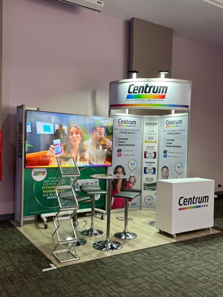 Stand promocional Centrum con pancartas, una pantalla de visualización de personas, un soporte para folletos, dos taburetes, una mesa y un mostrador con la marca en un entorno de conferencia o exposición.
