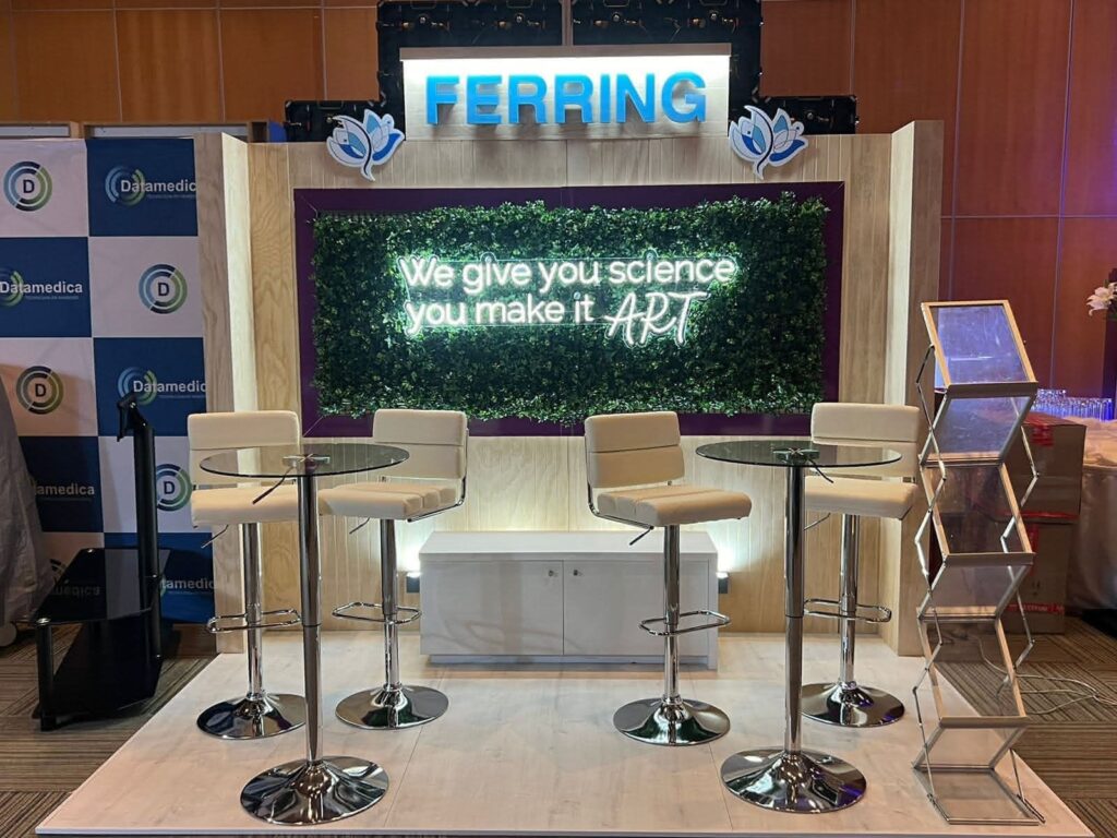 Una moderna caseta de exposición con el rótulo "Ferring" y una luz de neón que reza "Nosotros te damos ciencia, tú la conviertes en arte", con cuatro sillas blancas y dos mesas de cristal.