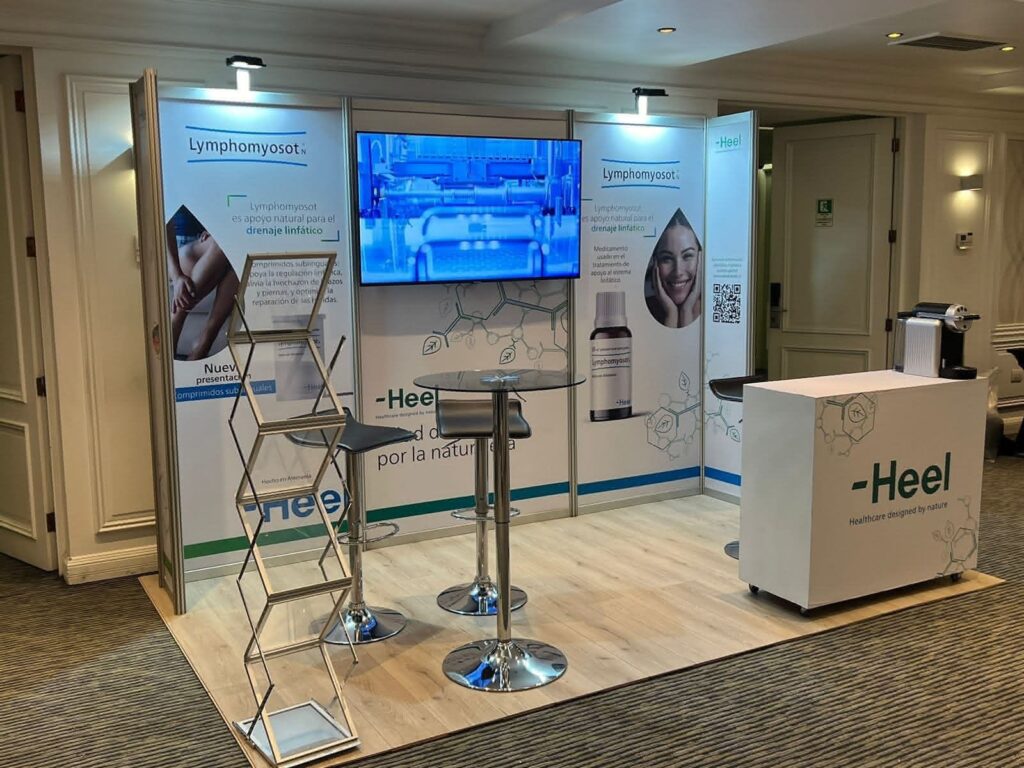 Stand de exposición para Heel con un expositor sobre Lymphomyosot, dos taburetes, un monitor, imágenes del producto y stands informativos en un entorno interior bien iluminado.