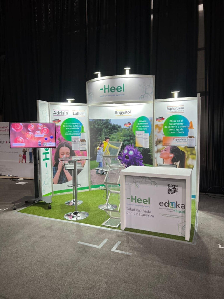 Stand de exposición para Heel con expositores de productos, pancartas, pantalla de televisión, sillas altas, mostrador con folletos y marca ecológica.