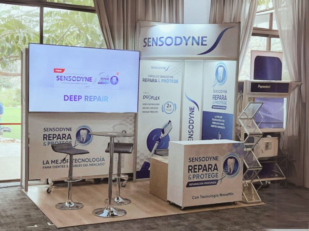 Stand promocional de Sensodyne con pancartas, pantalla de visualización, mesa alta con dos sillas y muestras de productos, instalado en el interior cerca de grandes ventanales.