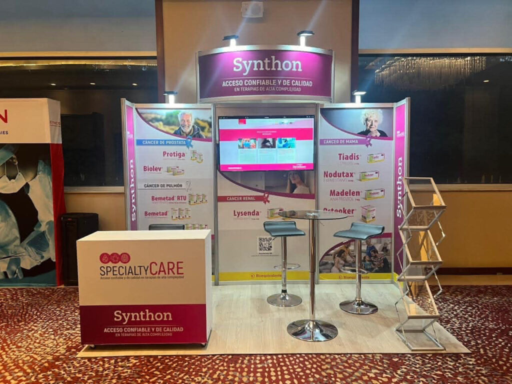 Stand de exposición para Synthon con información sobre los productos, dos taburetes, una mesa redonda, un monitor de visualización y un soporte para folletos, instalado sobre suelo enmoquetado.