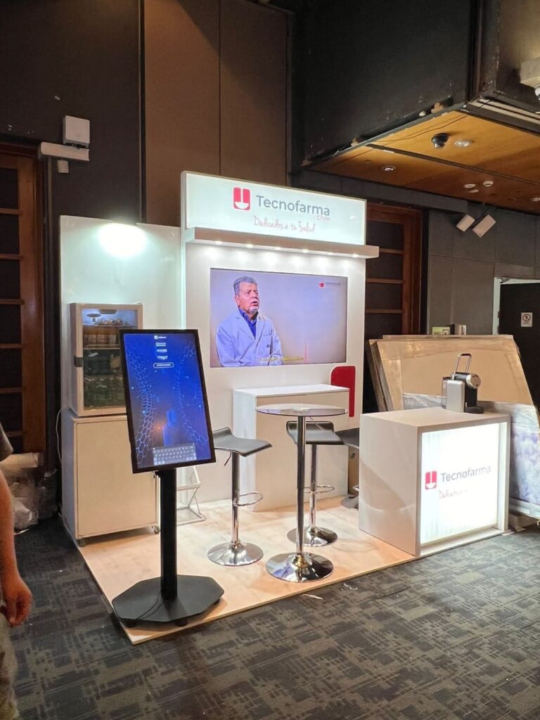 Stand de exposición de TecnoFarma con pantallas digitales, dos taburetes, señalización de marca y una pantalla de vídeo que muestra a un hombre hablando.