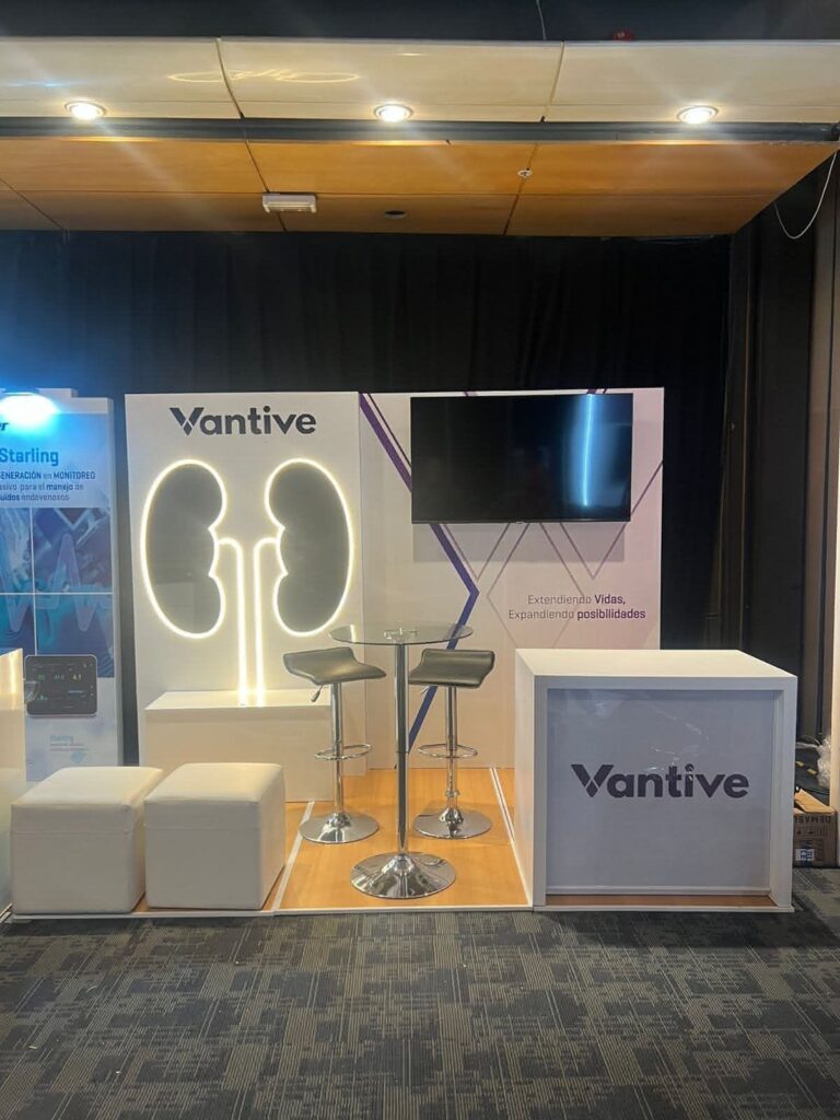 Stand de exposición con logotipo de Vantive, gráfico de riñón, dos taburetes, una mesa, un monitor grande y cubos blancos para sentarse sobre suelo de madera.
