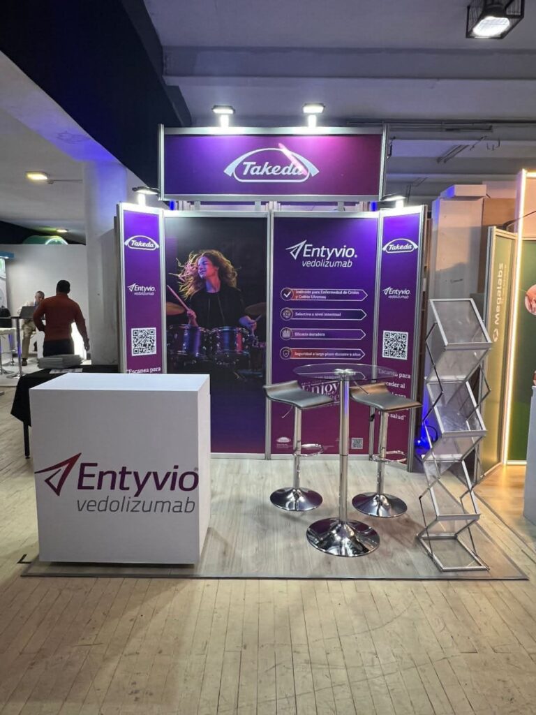Stand de Entyvio (vedolizumab), de Takeda, con señalización de marca, taburetes, mostrador y expositor en una feria de interior.