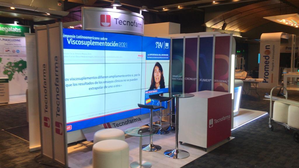 Stand de exposici&oacute;n de TecnoFarma con pantallas digitales que muestran diapositivas de presentaci&oacute;n y material promocional, rodeado de mobiliario de marca y paneles expositores en un evento en interiores.