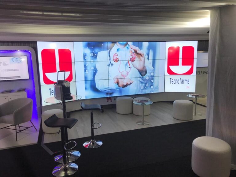 Un moderno stand de exposición con la marca TecnoFarma, pantallas digitales que muestran gráficos médicos, taburetes, sillas y pequeñas mesas dispuestas delante.