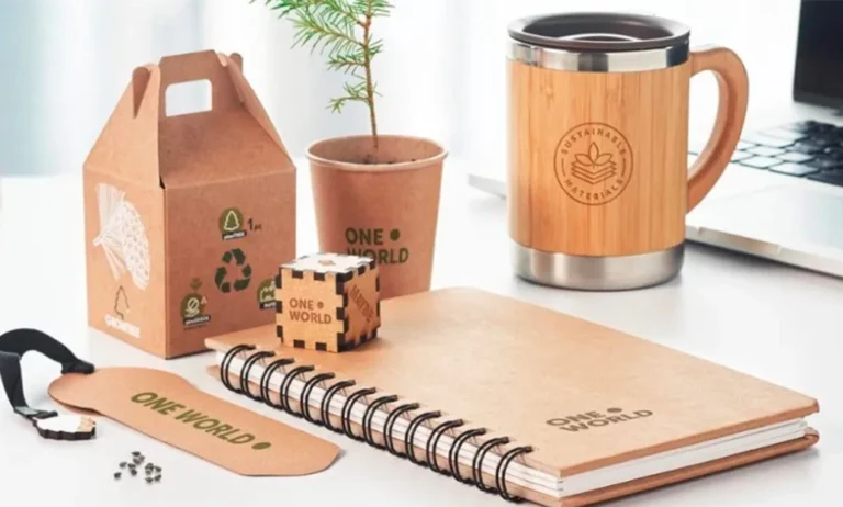 Una colección de artículos de oficina ecológicos que incluye un cuaderno, una taza, una pequeña planta en una maceta, un marcapáginas, un puzzle en forma de cubo y una caja de cartón, todo ello con la etiqueta "One World", sobre un escritorio.