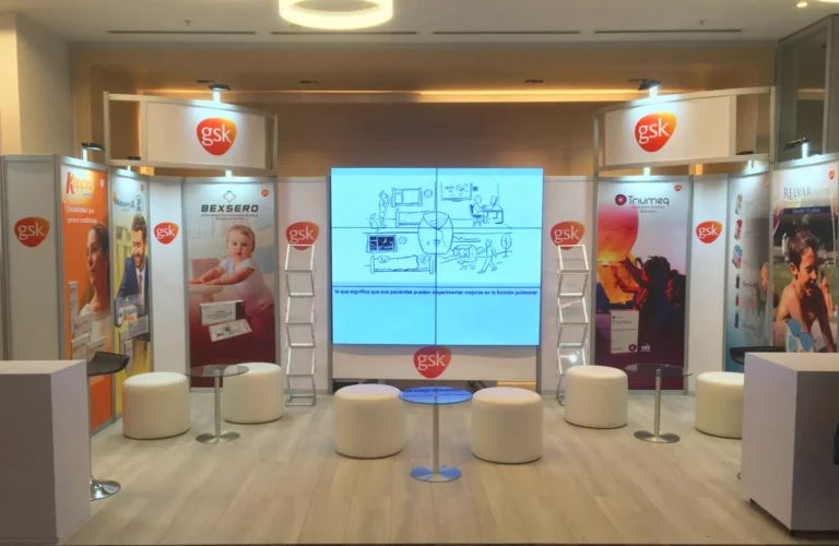 Stand de exposición de GSK con carteles promocionales, taburetes, mesas pequeñas y una pantalla central que muestra un diagrama, instalado en un espacio interior moderno.