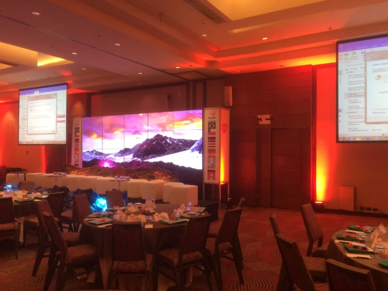 Una sala de conferencias con mesas redondas preparadas para un acto, grandes pantallas de visualización y un escenario con un paisaje montañoso en una pantalla central.