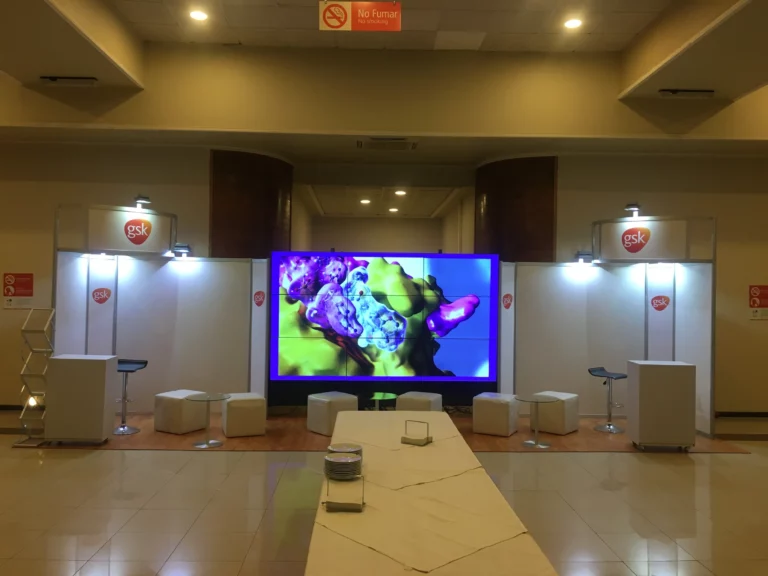 Stand de exposición con logotipos de GSK, cubos blancos para sentarse, una gran pantalla digital con formas abstractas y carteles "No Fumar" en un espacio interior muy iluminado.