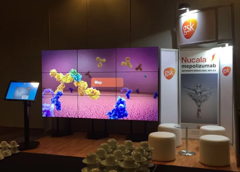 Stand farmacéutico con una gran pantalla que muestra un gráfico molecular, la marca Nucala, logotipos de GSK, zona de asientos y tazas apiladas en primer plano.