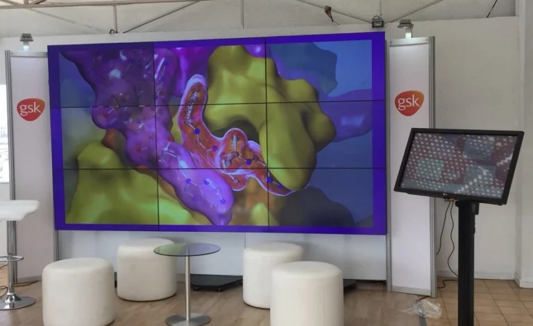 Una gran pantalla muestra un gráfico científico molecular frente a unos taburetes blancos y una mesa de cristal; a ambos lados se ven los logotipos de GSK y a la derecha hay una pantalla táctil.