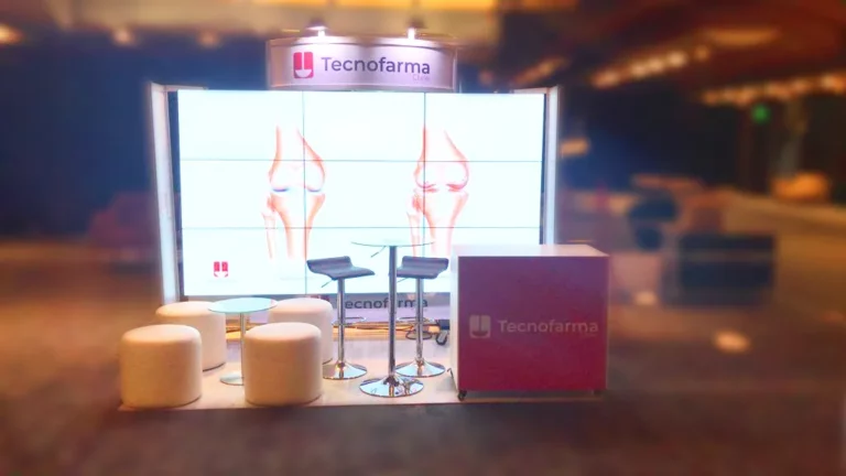 Un stand de exposición de TecnoFarma con taburetes, otomanas redondas, un mostrador y una pantalla que muestra imágenes médicas de articulaciones de rodilla.