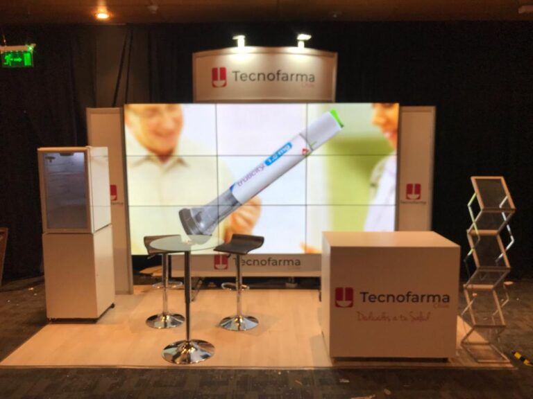 Stand de exposición de TecnoFarma con un mostrador, dos taburetes, una pantalla que muestra un dispositivo médico, un soporte para folletos y marcas en las paredes y el mobiliario.