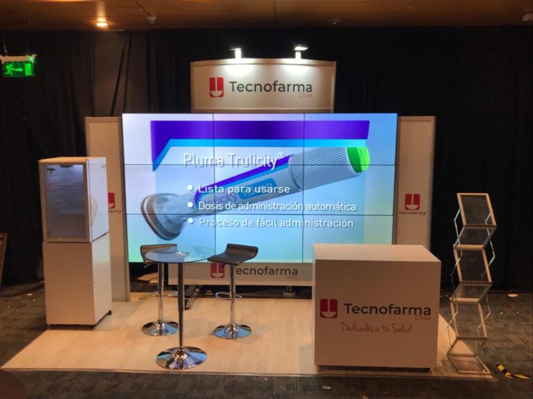 Stand de exposición de TecnoFarma con paneles de marca, una pantalla que muestra información sobre Trulicity, dos sillas, una mesa, un soporte para folletos y unidades de almacenamiento.