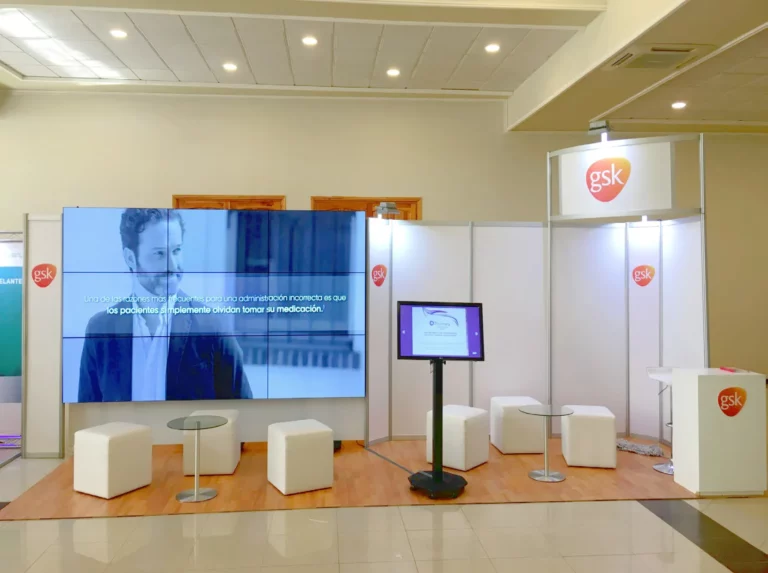 Stand de exposición de GSK con pantalla digital que muestra a un hombre y texto en español, taburetes blancos, mesas pequeñas y logotipos de la empresa en las paredes y el mostrador.