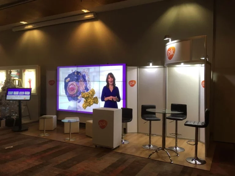 Stand de exposición con la marca GSK, una gran pantalla de vídeo que muestra a un presentador, tres taburetes en un mostrador y una pequeña zona de asientos.