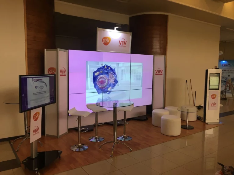 Stand de exposición con una gran pantalla digital que muestra una imagen científica, con los logotipos de GSK y ViiV, rodeada de taburetes, un monitor táctil y pantallas informativas.