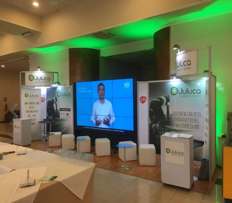 Stand de la conferencia sobre el tratamiento del VIH Juluca con pancartas, asientos, iluminación verde y una gran pantalla en la que aparece un hombre haciendo una presentación.