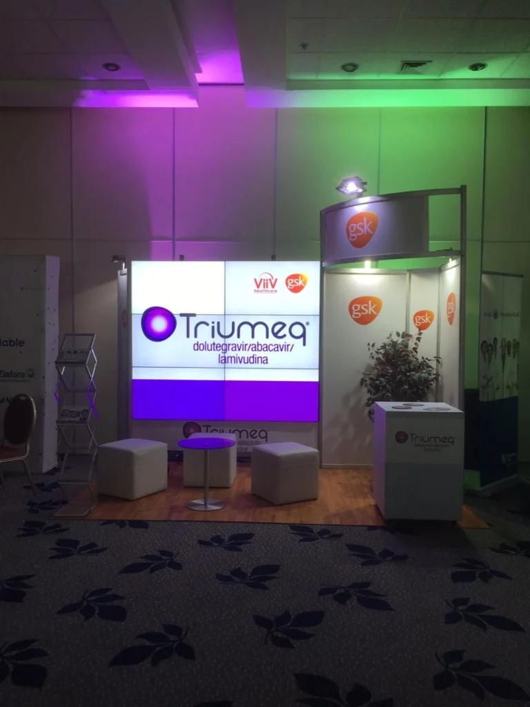Stand de exposición para Triumeq de ViiV Healthcare y GSK, con señalización de marca, taburetes para sentarse y un pequeño mostrador con una planta.