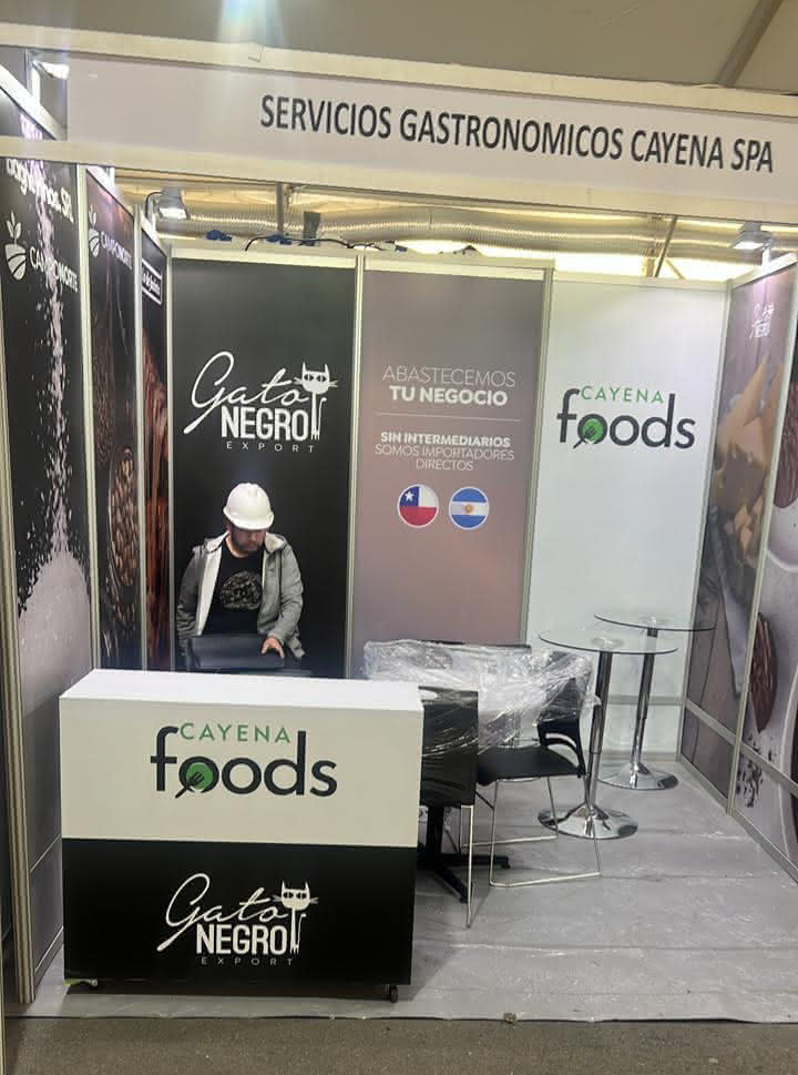 Un hombre con casco se sienta en un stand rotulado "Cayena Foods" y "Gato Negro Export" en una feria de Servicios Gastronómicos Cayena SPA.