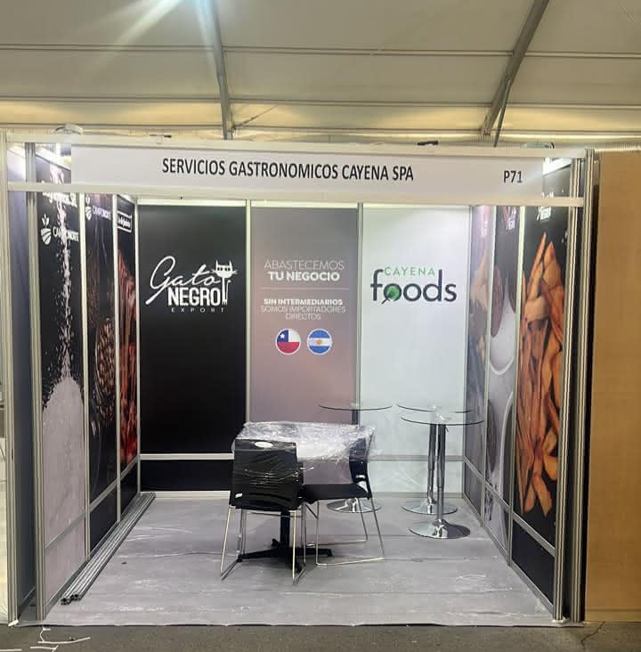 Stand de feria para Servicios Gastronómicos Cayena SPA con la marca Gato Negro Export y Cayena Foods, con una mesa, sillas e imágenes de alimentos en paneles.