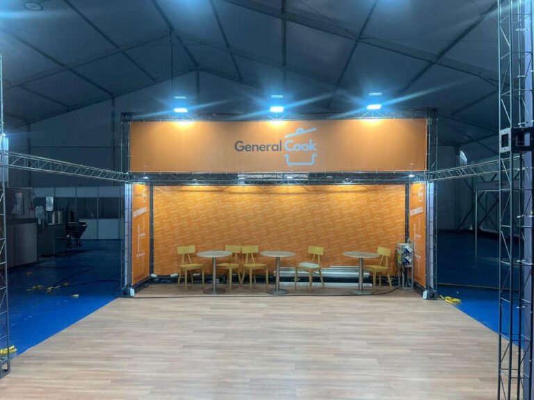 Un stand de exposición con la marca "General Cook", con paredes naranjas, mesas y sillas de madera, instalado dentro de una gran carpa con suelo azul.