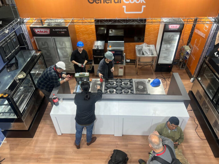 Cinco personas instalan electrodomésticos y equipos en una sala de exposición de cocinas o en un stand de una feria comercial, con hornos, fogones y carteles de exposición al fondo.