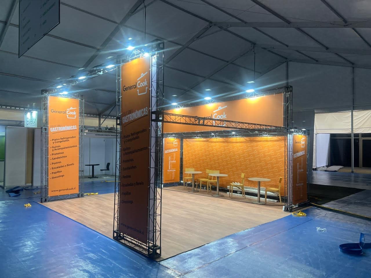 Stand de exposición naranja con la marca "General Cook", carteles luminosos, suelo de madera y mesas con taburetes, instalado en un gran recinto interior con suelo azul.