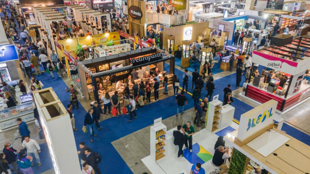 La multitud camina entre los stands y expositores de una concurrida feria o exposición de interior, con pancartas y productos visibles por todo el recinto.