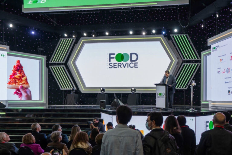 Un orador sube a un podio en el escenario de un evento de servicios alimentarios, con una gran pantalla que muestra "FOOD SERVICE" y otra pantalla que muestra una mano que sostiene una porción de pizza.