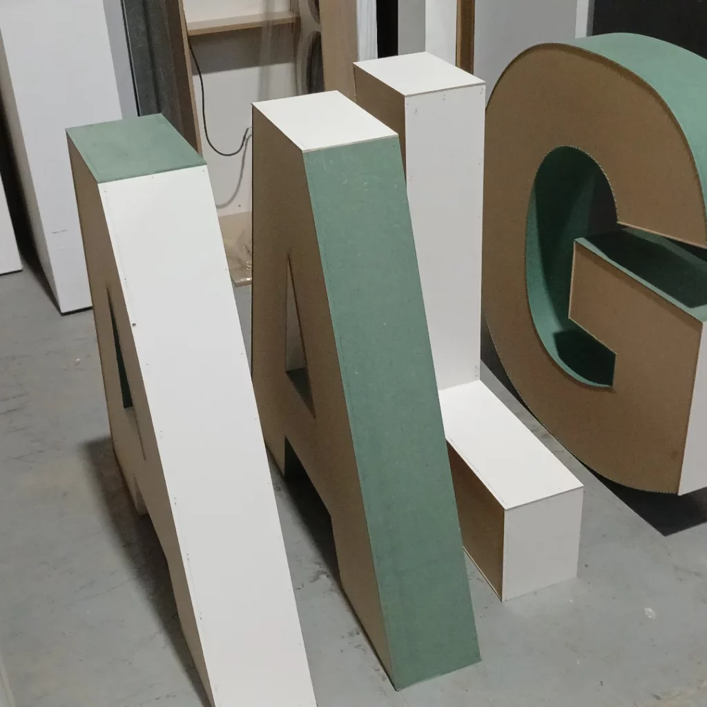 Grandes letras tridimensionales "A", "I" y "G" de madera o material similar, erguidas sobre el suelo de un taller.
