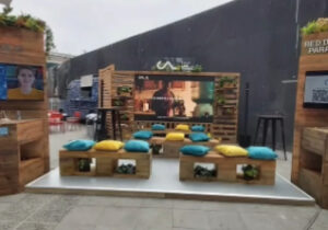 Montaje de un evento al aire libre con bancos de madera, cojines de colores, una gran pantalla y plantas decorativas, dispuestos en una plataforma frente a una pared oscura de fondo.