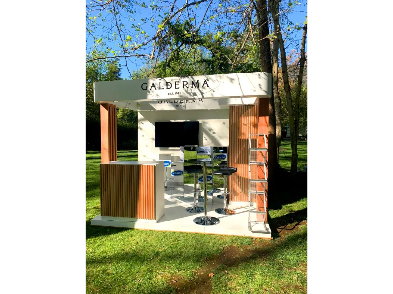 Stand promocional al aire libre para Galderma con asientos, mostrador, pantalla de TV y estanterías de exposición, situado sobre césped verde entre árboles a la luz del día.